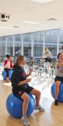 students sit on exercise balls tossing a yellow ball. panel of windows behind them